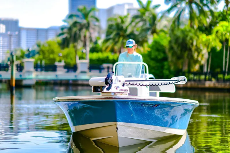 Slide: The Image of 2022 Hewes Redfisher 21 boat on calm water, with lush greenery and cityscape background. - 7