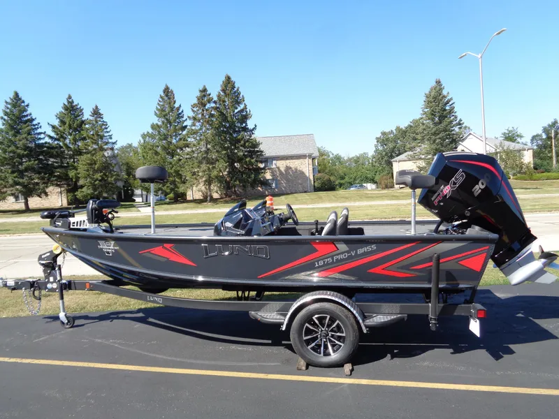 The Image of 2025 Lund 1875 Pro-V Bass XS boat on trailer, black with red accents, parked outdoors. - 0