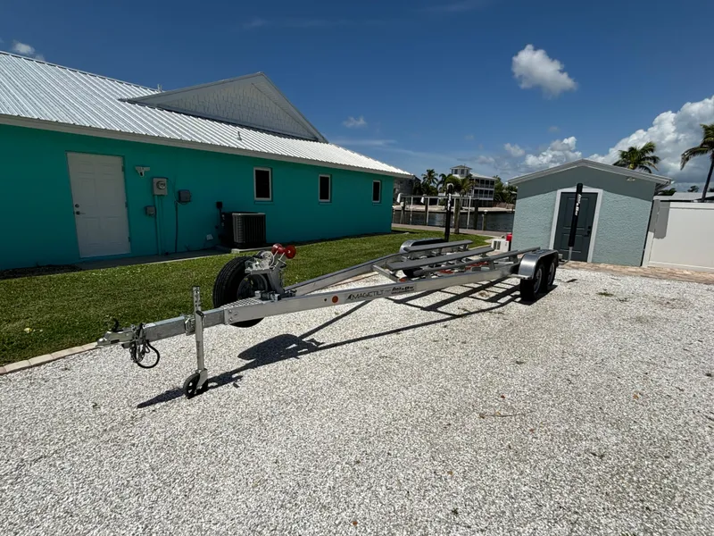 Slide: The Image of 2024 Lake & Bay 25 Open boat trailer parked on gravel near turquoise building. - 22