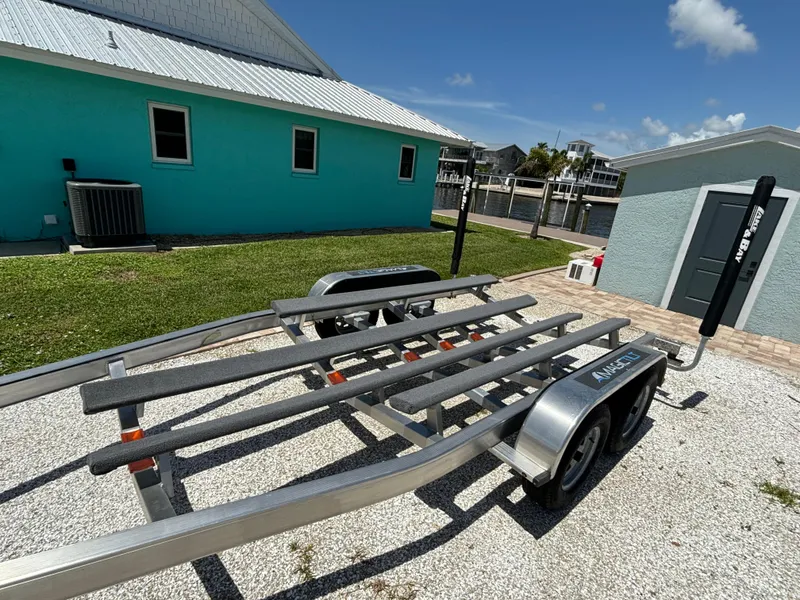 Slide: The Image of 2024 Lake & Bay 25 Open boat trailer parked on gravel near turquoise building. - 23