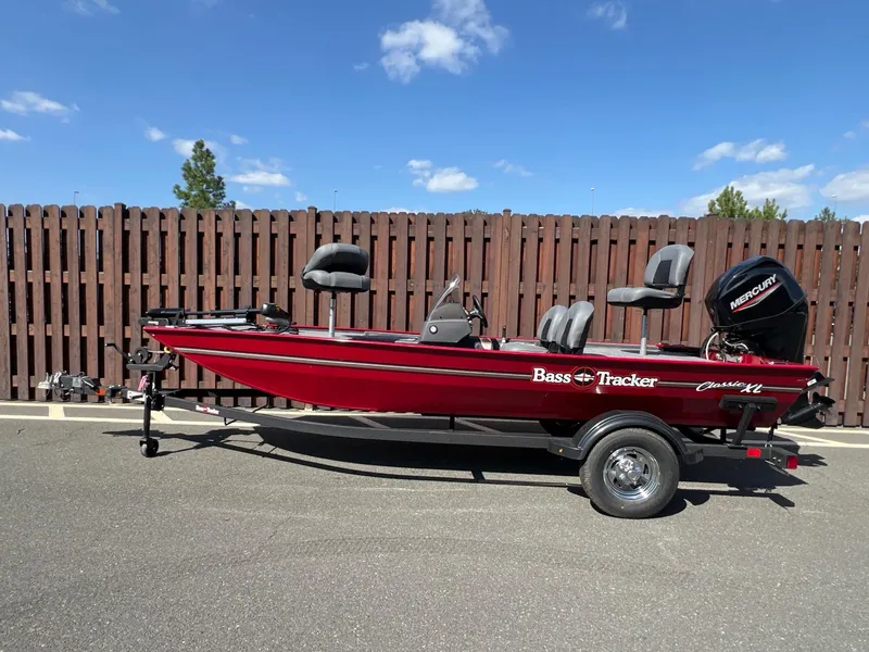 The Image of 2025 Tracker Bass Tracker Classic XL boat, red, parked on trailer, wooden fence background. - 0