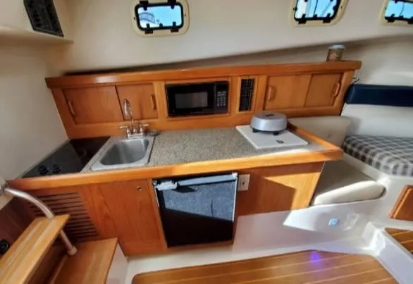 Slide: The Image of 1999 Mainship 30 interior kitchen with wooden cabinets, sink, microwave, and seating area. - 19