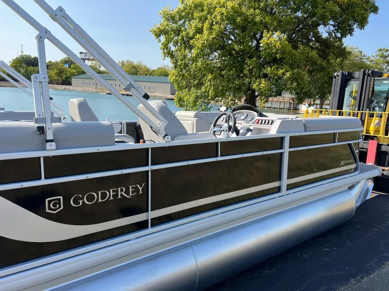 Slide: The Image of 2025 Godfrey Xperience 2286 SBX pontoon boat docked by a scenic waterfront. - 2