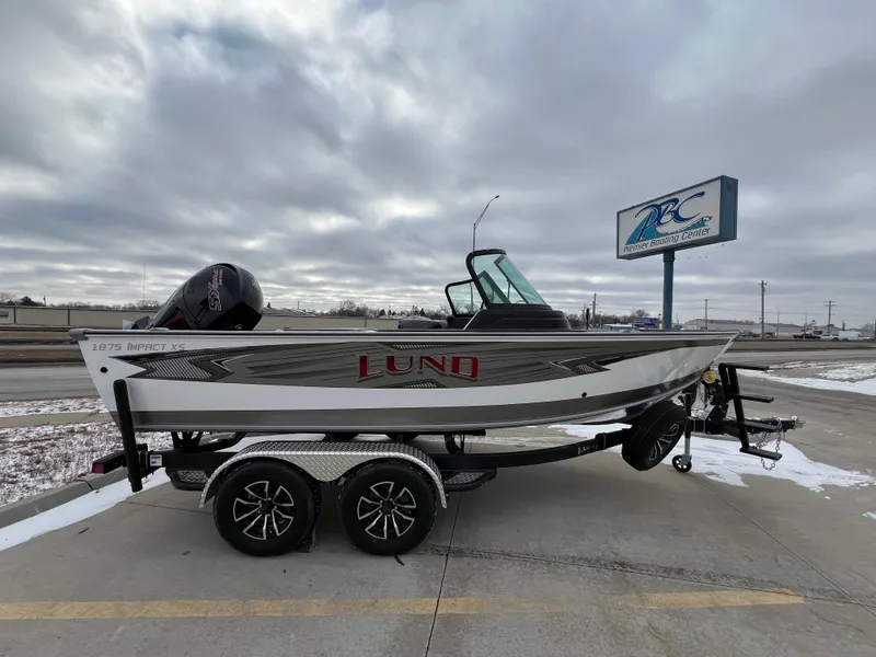Slide: The Image of 2025 Lund 1875 Impact XS Sport boat on trailer, parked outdoors under cloudy sky. - 0