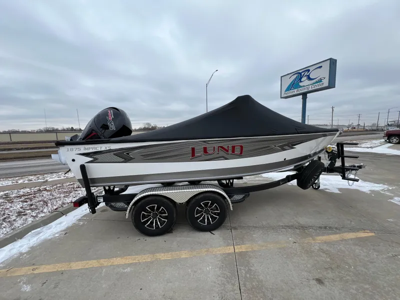 Slide: The Image of 2025 Lund 1875 Impact XS Sport boat on trailer, covered, parked in snowy lot. - 2