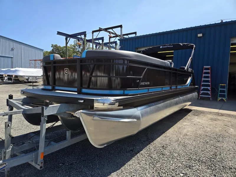 The Image of 2025 Godfrey Sweetwater 2486 SB pontoon boat on trailer, parked outside blue warehouse. - 0