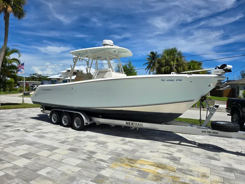 The Image of 2015 Cobia 296 Center Console boat on trailer, parked in a driveway under a clear blue sky. - 0