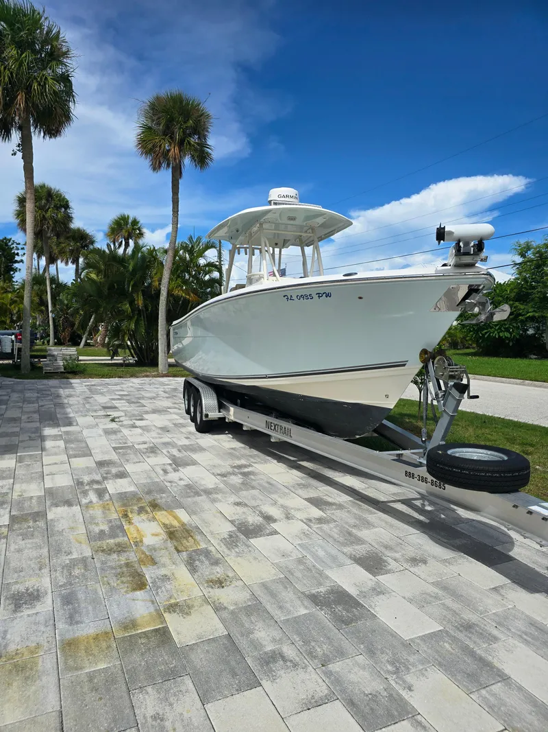 Slide: The Image of 2015 Cobia 296 Center Console boat on trailer, parked on paved driveway, surrounded by palm trees. - 49