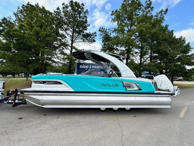 Slide: The Image of 2024 Avalon Quad Lounge Windshield boat, turquoise and silver, parked outdoors with trees in background. - 1