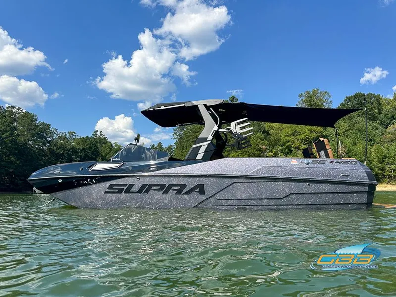The Image of 2024 Supra SE boat on a lake, under a clear blue sky with trees in the background. - 0