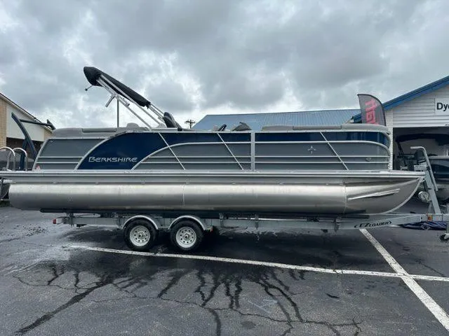 Slide: The Image of 2023 Berkshire 24ULE-STS3 pontoon boat on trailer, parked outdoors under cloudy sky. - 2