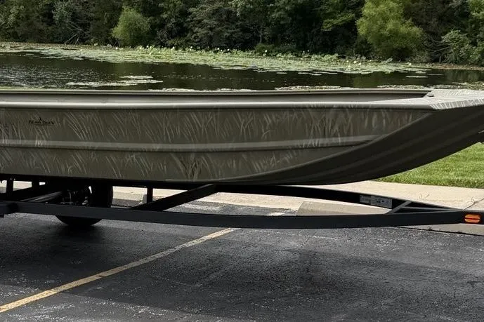 The Image of 2025 Tracker Grizzly 1754 Jon boat on trailer by a lake with lush greenery. - 0
