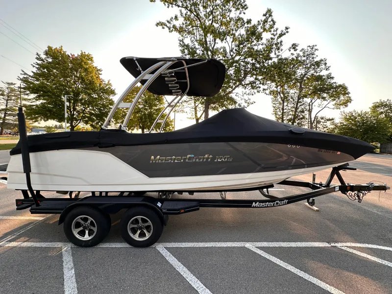 Slide: The Image of 2017 MasterCraft NXT22 boat on trailer, parked in a lot with trees in the background. - 18