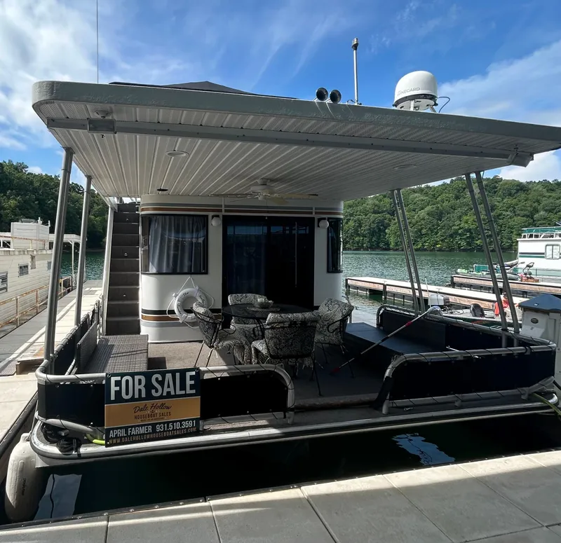 Slide: The Image of 1999 Lakeview houseboat for sale, docked on a scenic lake with outdoor seating. - 2