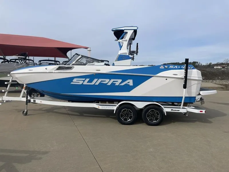 Slide: The Image of 2023 Supra SA 450 boat on trailer, blue and white design, parked outdoors. - 1