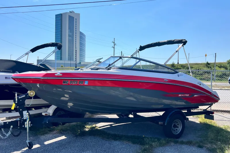 Slide: The Image of 2021 Yamaha Boats SX195 on trailer, red and gray, parked outdoors near a tall building. - 2