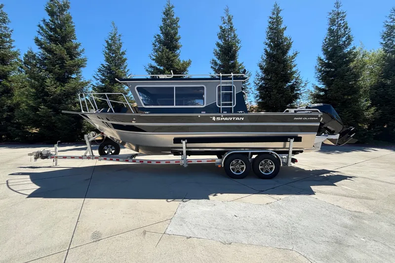 The Image of 2025 Spartan 2600 Olympia boat on trailer, surrounded by trees, under clear blue sky. - 0