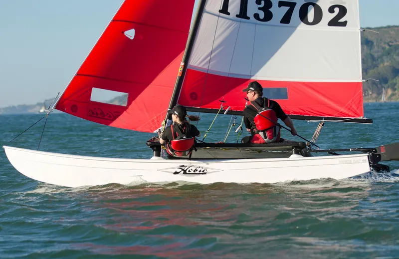 Slide: The Image of 2025 Hobie Cat 16 Catamaran sailing with red sails on open water. - 3