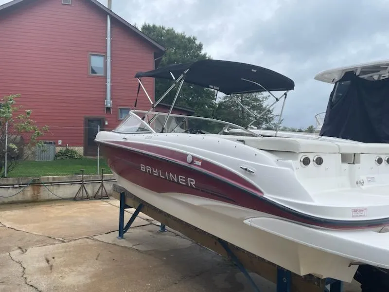 Slide: The Image of 2011 Bayliner 235 Bowrider boat with canopy, parked near a red house. - 2