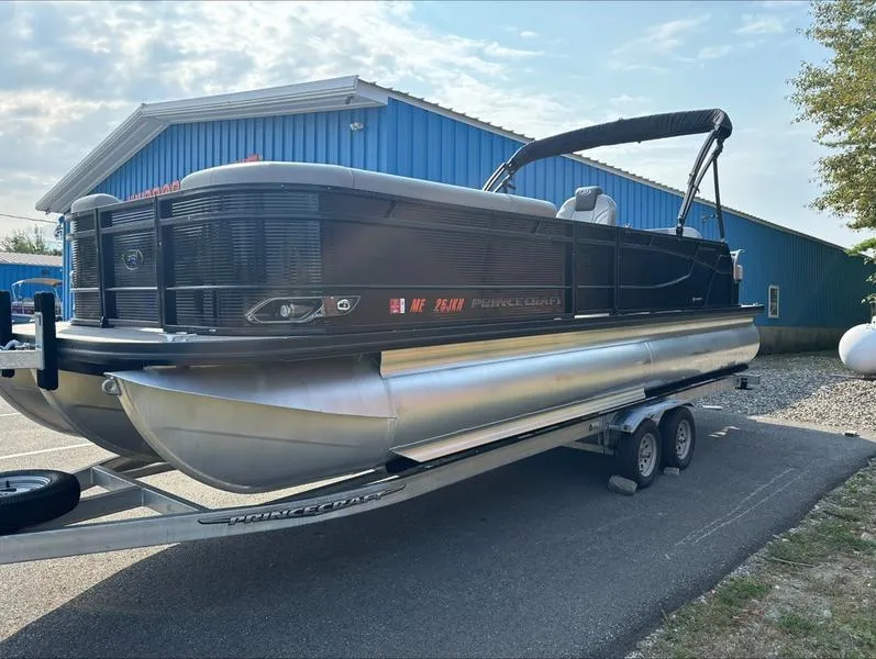 Slide: The Image of 2023 Princecraft QUORUM Q25 pontoon boat on trailer, parked near blue building. - 0