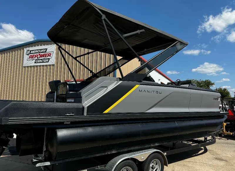 Slide: The Image of 2024 Manitou Explore pontoon boat with canopy, parked on a trailer outside a repower center. - 3