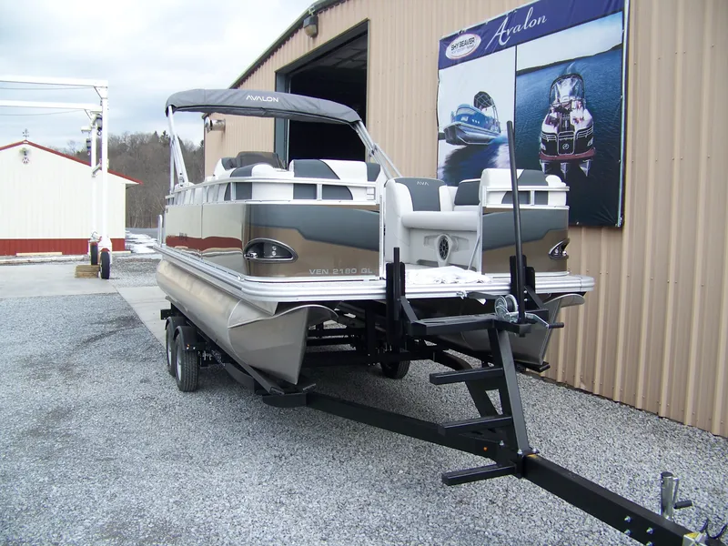 Slide: The Image of 2025 Avalon Venture 21QL pontoon boat on trailer outside a warehouse. - 2