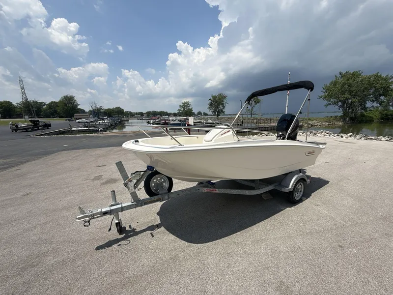 Slide: The Image of 2025 Boston Whaler 130 Super Sport boat on trailer, parked near a lake under cloudy sky. - 2