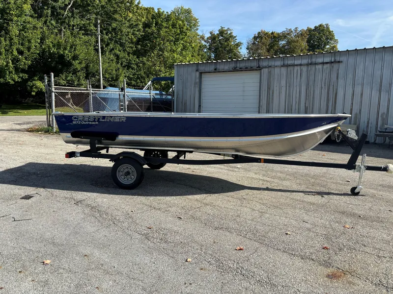 The Image of 2025 Crestliner 1672 Outlook boat on trailer, parked outdoors near a metal building. - 0