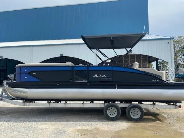 Slide: The Image of 2023 Barletta Cabrio 24 UC pontoon boat on trailer, parked near a blue building. - 0