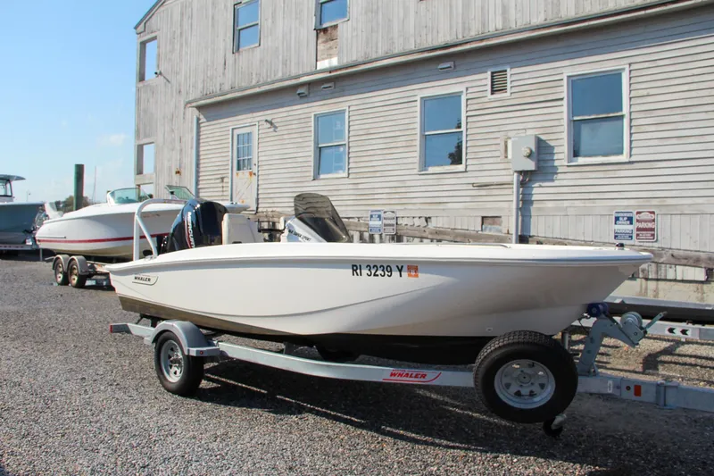 Slide: The Image of 2022 Boston Whaler 160 Super Sport boat on trailer, parked near a wooden building. - 22