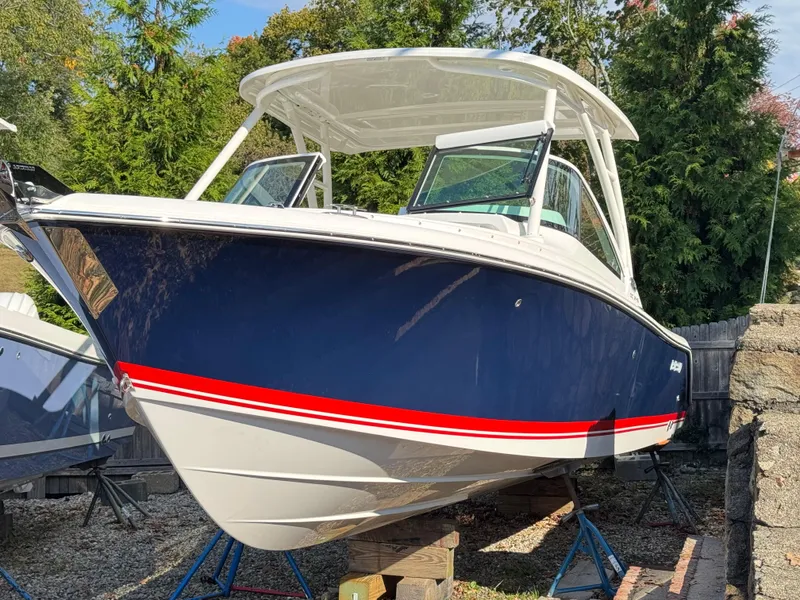 The Image of 2025 Pursuit DC 246 Dual Console boat with blue hull and red stripe, displayed outdoors. - 0