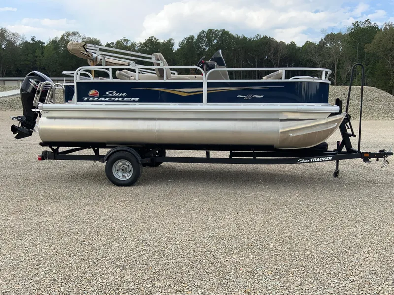The Image of 2025 Sun Tracker Bass Buggy 18 DLX pontoon boat on trailer, parked on gravel. - 1