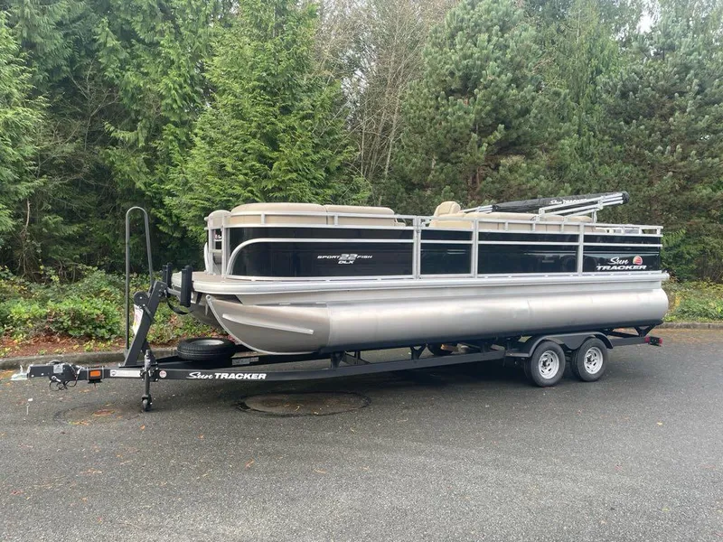 The Image of 2025 Sun Tracker SportFish 22 DLX pontoon boat on trailer, parked near lush greenery. - 0