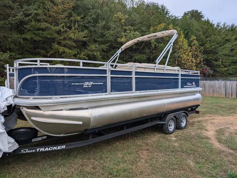 The Image of 2025 Sun Tracker Fishin' Barge 22 DLX pontoon boat on trailer, parked on grass. - 0