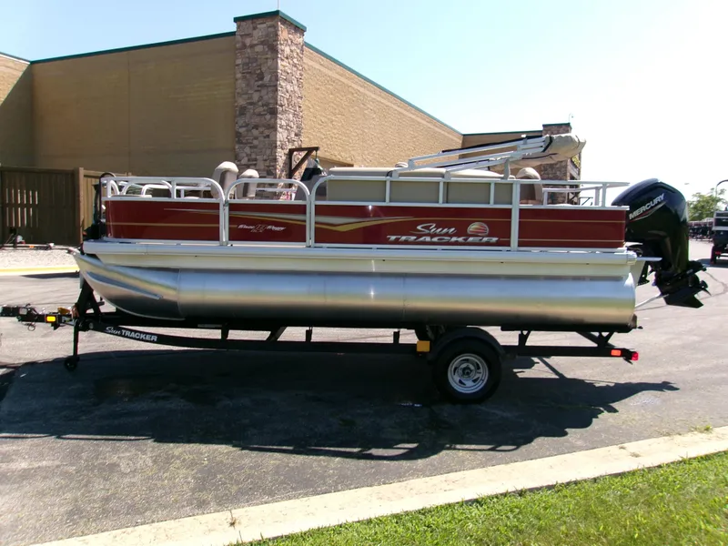 Slide: The Image of 2025 Sun Tracker Bass Buggy 18 DLX pontoon boat on trailer, parked outdoors. - 2