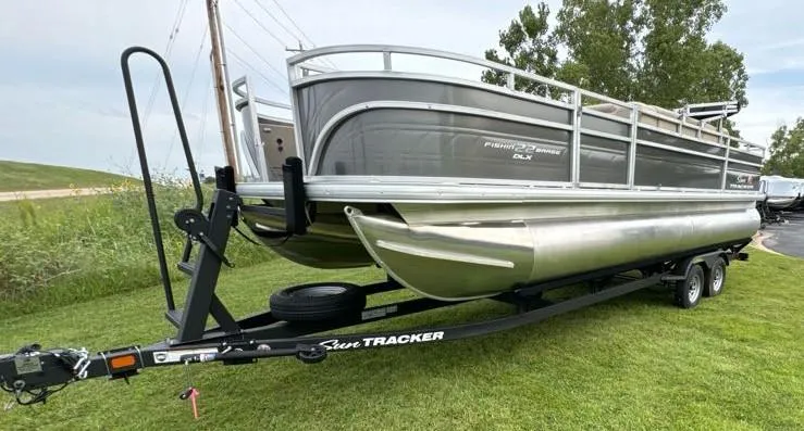 Slide: The Image of 2025 Sun Tracker Fishin' Barge 22 DLX pontoon boat on trailer, parked on grass. - 2