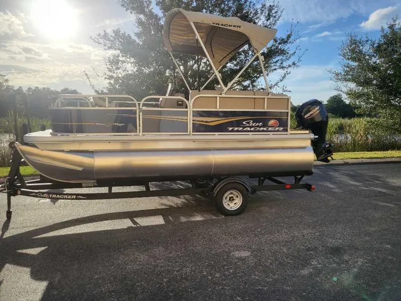 Slide: The Image of 2025 Sun Tracker Bass Buggy 16 XL Select pontoon boat on trailer, sunny day. - 2
