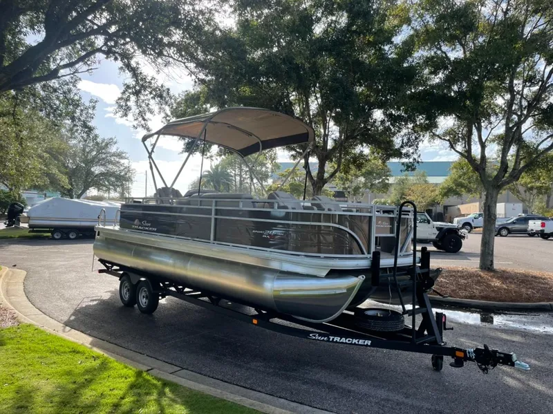 Slide: The Image of 2025 Sun Tracker Sportfish 24 XP3 pontoon boat on trailer, parked under trees. - 2