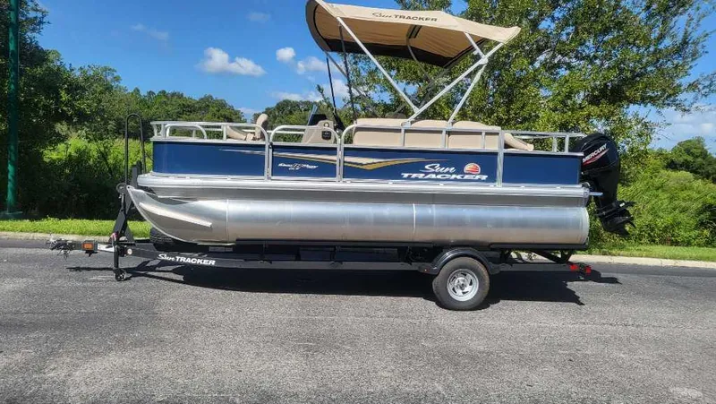 Slide: The Image of 2025 Sun Tracker Bass Buggy 18 DLX pontoon boat on trailer, parked outdoors. - 2