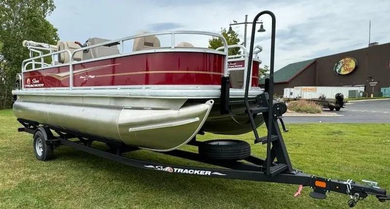 Slide: The Image of 2025 Sun Tracker Bass Buggy 18 DLX pontoon boat on trailer, parked on grass. - 2