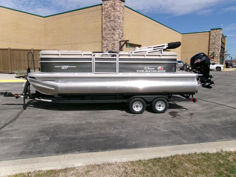 Slide: The Image of 2025 Sun Tracker Party Barge 20 DLX pontoon boat on trailer, parked outdoors. - 2