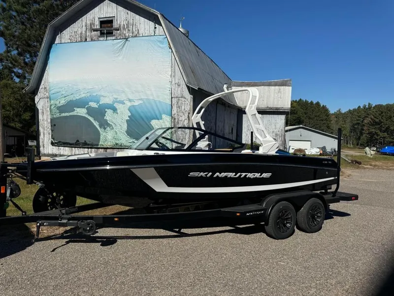 Slide: The Image of 2025 Nautique Ski 200 Open Bow boat on trailer, parked near rustic barn. - 9