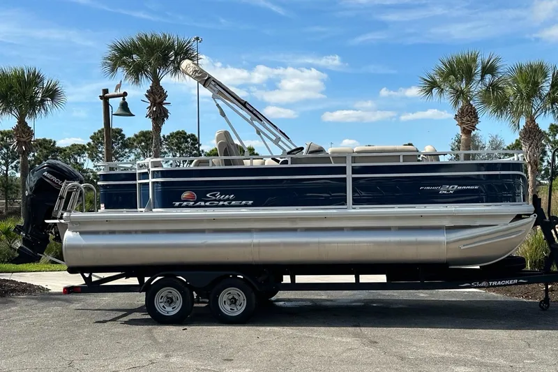 Slide: The Image of 2023 Sun Tracker Fishin' Barge 20 DLX pontoon boat on trailer, parked near palm trees. - 2
