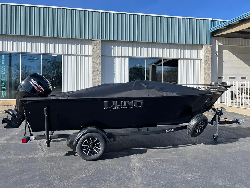 Slide: The Image of 2025 Lund 1650 Rebel XL SS boat with cover, parked outside a storage facility. - 26