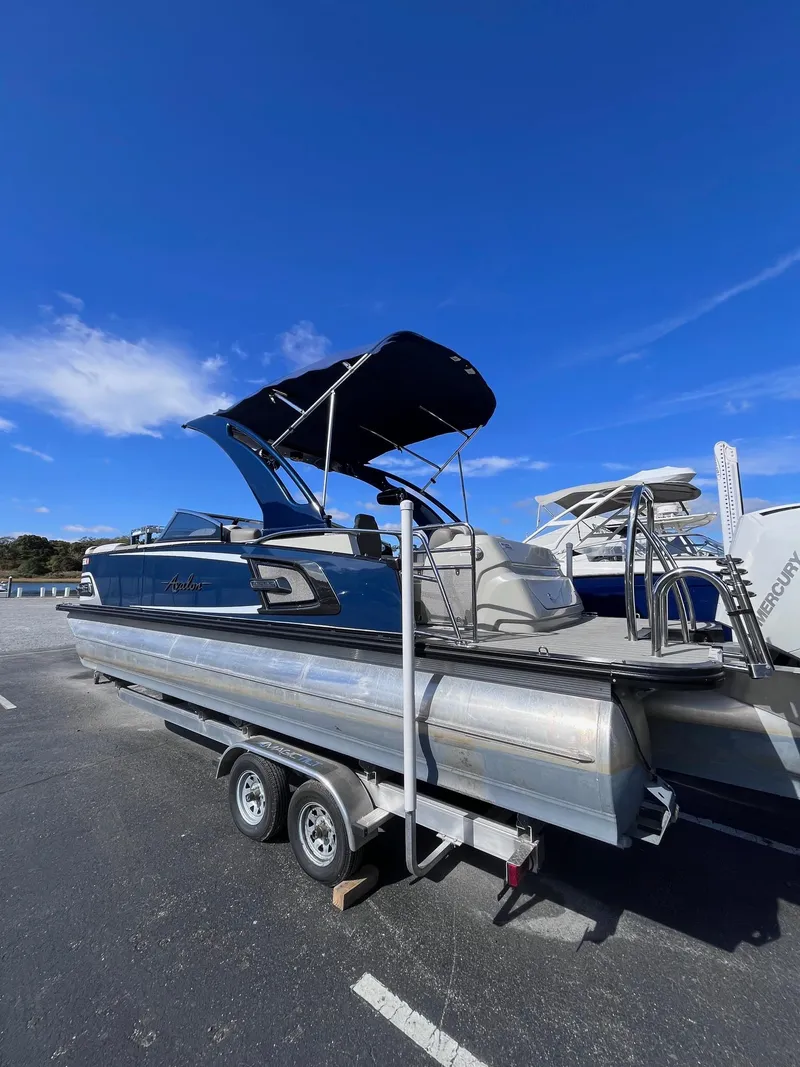 Slide: The Image of 2023 Avalon Excalibur Elite Windshield boat on trailer under clear blue sky. - 2