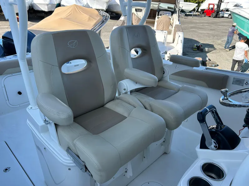 Slide: The Image of 2018 Sailfish 290 CC boat seats, beige upholstery, armrests, and cup holders in a marina setting. - 11