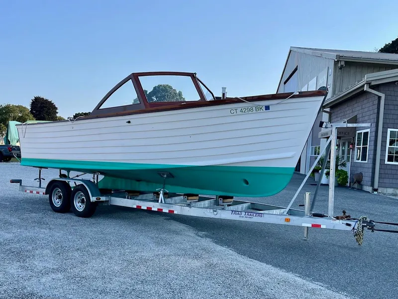 Slide: The Image of 2024 MacKenzie Cuttyhunk boat on trailer, white and turquoise, parked outside a building. - 49