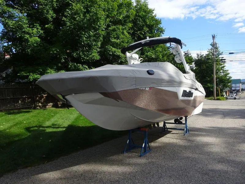 Slide: The Image of 2020 Malibu M240 boat on stands, parked on a driveway with green trees. - 2