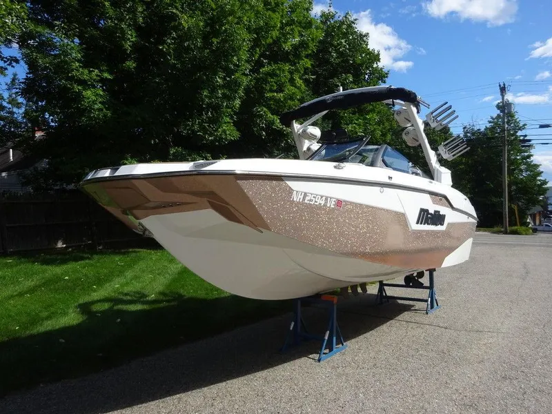 Slide: The Image of 2020 Malibu M240 boat on stands, parked on a driveway with trees in the background. - 5