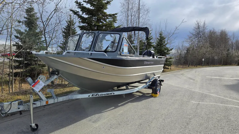 Slide: The Image of 2024 Spartan 185 Astoria boat on trailer, parked on a road with trees in the background. - 2